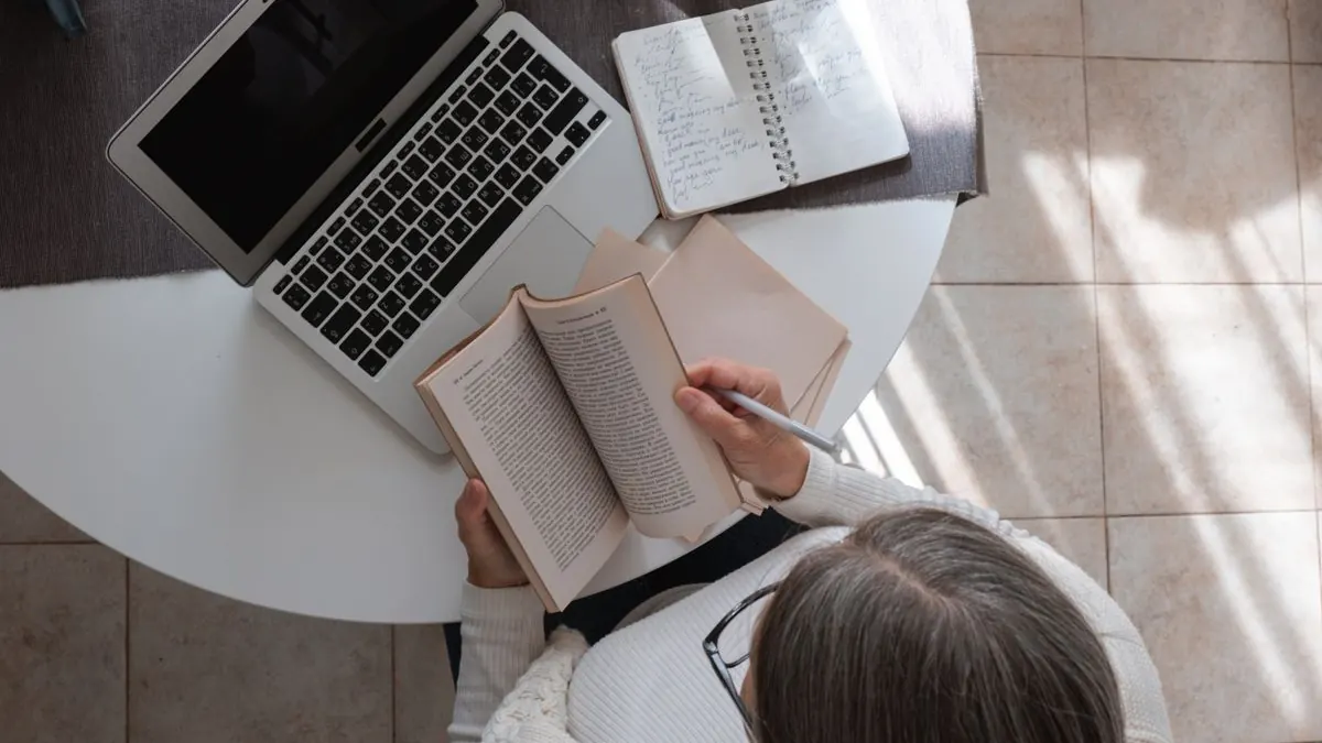 Žena čte knihu vedle notebooku na stole